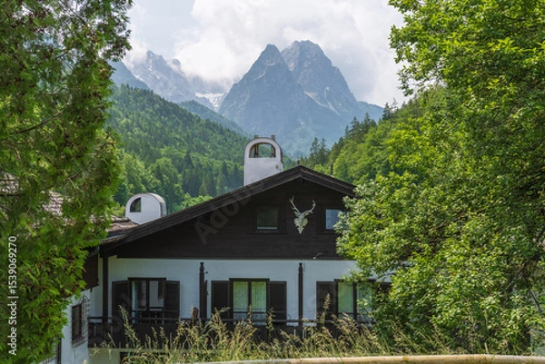Fototapeta Charming alpine buildings in Mittenwald feature colorful Lüftlmalerei (frescoes), wooden balconies, and flower-filled window boxes, all set against a dramatic mountain backdrop.