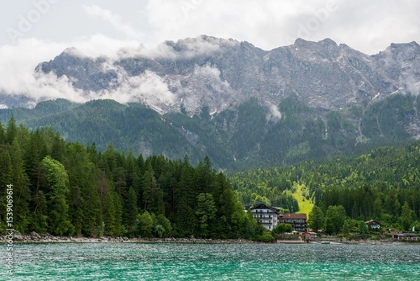 Fototapeta A serene alpine lake with crystal-clear turquoise water mirrors the snow-capped Zugspitze and pine-covered hills. Rowboats glide gently under a clear blue sky—pure alpine tranquility.