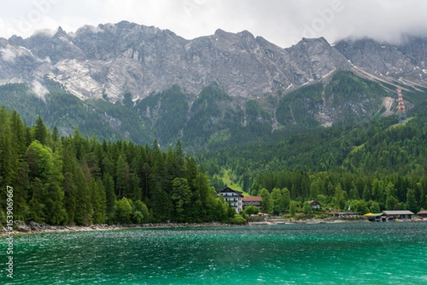 Fototapeta A serene alpine lake with crystal-clear turquoise water mirrors the snow-capped Zugspitze and pine-covered hills. Rowboats glide gently under a clear blue sky—pure alpine tranquility.