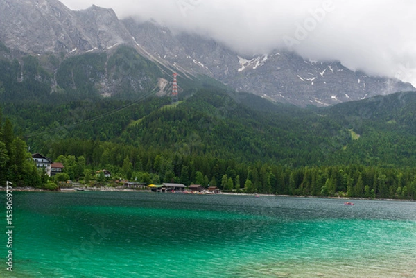 Fototapeta A serene alpine lake with crystal-clear turquoise water mirrors the snow-capped Zugspitze and pine-covered hills. Rowboats glide gently under a clear blue sky—pure alpine tranquility.