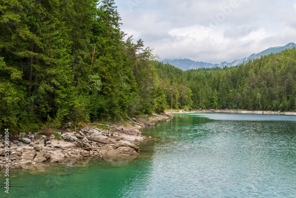 Fototapeta A serene alpine lake with crystal-clear turquoise water mirrors the snow-capped Zugspitze and pine-covered hills. Rowboats glide gently under a clear blue sky—pure alpine tranquility.