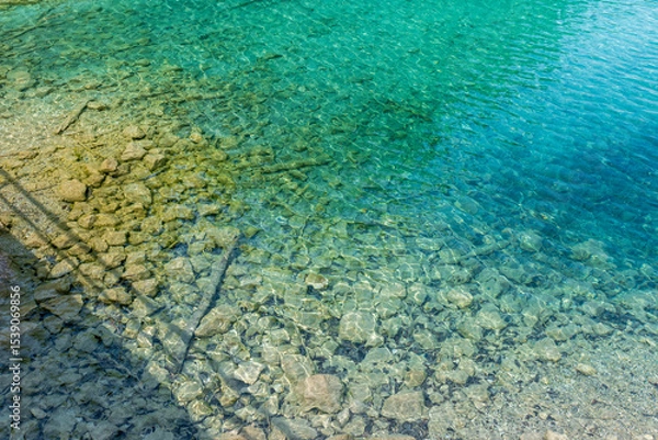 Fototapeta A serene alpine lake with crystal-clear turquoise water mirrors the snow-capped Zugspitze and pine-covered hills. Rowboats glide gently under a clear blue sky—pure alpine tranquility.