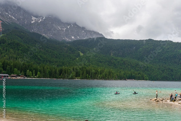 Fototapeta A serene alpine lake with crystal-clear turquoise water mirrors the snow-capped Zugspitze and pine-covered hills. Rowboats glide gently under a clear blue sky—pure alpine tranquility.