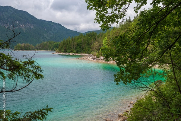 Fototapeta A serene alpine lake with crystal-clear turquoise water mirrors the snow-capped Zugspitze and pine-covered hills. Rowboats glide gently under a clear blue sky—pure alpine tranquility.