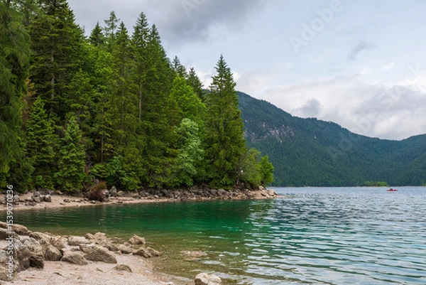 Fototapeta A serene alpine lake with crystal-clear turquoise water mirrors the snow-capped Zugspitze and pine-covered hills. Rowboats glide gently under a clear blue sky—pure alpine tranquility.