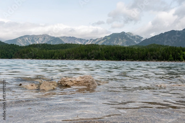 Fototapeta A serene alpine lake with crystal-clear turquoise water mirrors the snow-capped Zugspitze and pine-covered hills. Rowboats glide gently under a clear blue sky—pure alpine tranquility.