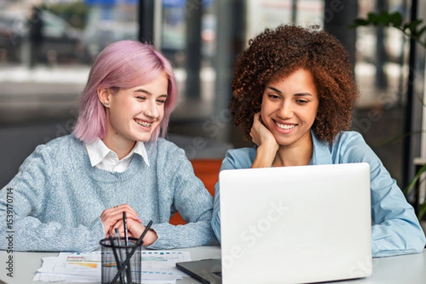 Fototapeta Smiling diverse young women collaborating on laptop in modern coworking space. Concept of teamwork, digital nomads, freelance, generation Z and creative business