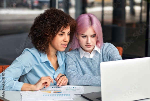 Fototapeta Young diverse women working together at laptop and analyzing business documents. Concept of teamwork, digital collaboration, mentorship and modern workplace