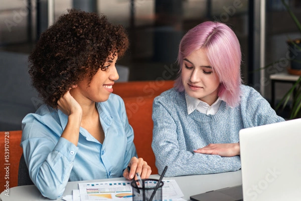 Fototapeta Young diverse women working together at laptop and analyzing business documents. Concept of teamwork, digital collaboration, mentorship and modern workplace
