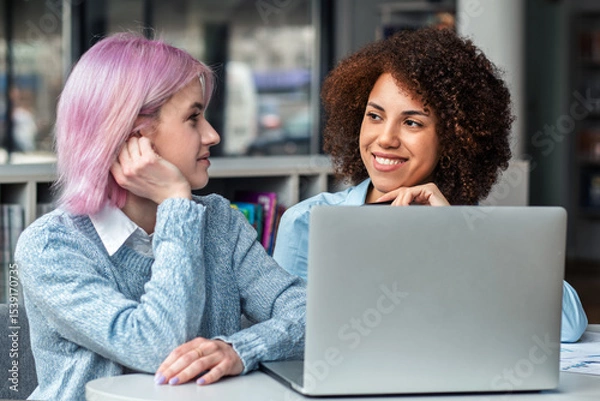 Fototapeta Smiling diverse young women collaborating on laptop in modern coworking space. Concept of teamwork, digital nomads, freelance, generation Z and creative business