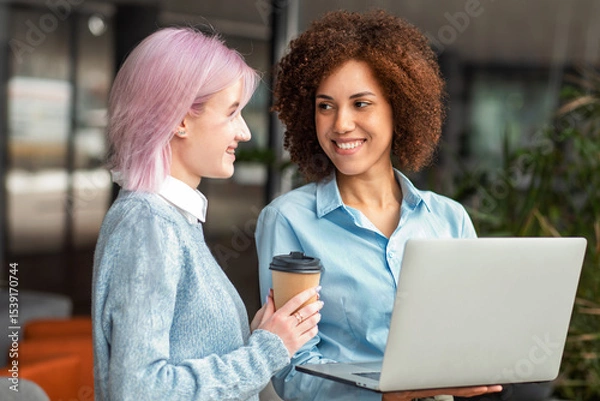 Fototapeta Two smiling young women having friendly conversation while holding laptop and coffee in modern workspace. Concept of networking, teamwork and female empowerment