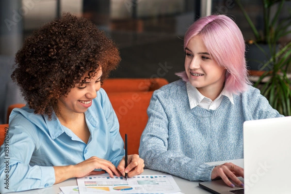 Fototapeta Smiling diverse young women collaborating on laptop in modern coworking space. Concept of teamwork, digital nomads, freelance, generation Z and creative business