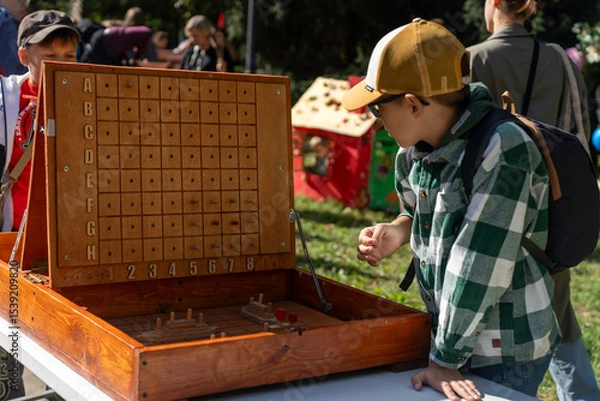 Obraz Curious boy in checkered shirt and cap playing large wooden battleship game outdoors on sunny day during family festival. Concept of childhood fun, outdoor games, learning through play and strategy.