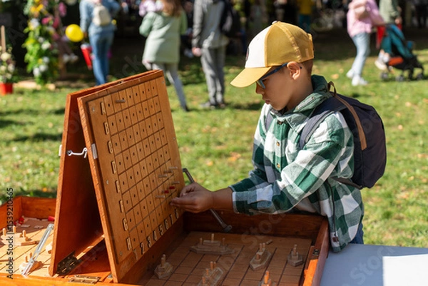 Obraz Boy wearing plaid shirt and baseball cap concentrates on playing giant wooden board game outdoors during public event. Concept of educational entertainment, strategic thinking, and childhood joy.