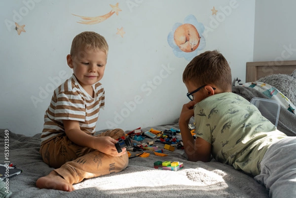 Obraz Two happy little boys playing with colorful plastic construction bricks on bed in cozy children room with wall decals. Concept of childhood, creativity, indoor play and sibling bonding.
