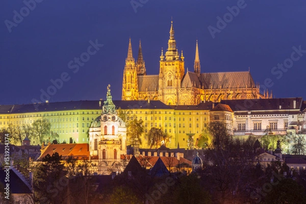 Fototapeta Illuminated Saint Vitus Cathedral and surrounding buildings in Prague Czech Republic at night with gothic towers and Baroque church in foreground
