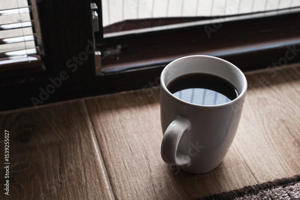 Obraz Minimalistic morning scene with a cup of black coffee on a wooden floor near a window, soft sunlight and shadows creating cozy, peaceful atmosphere and warm tones
