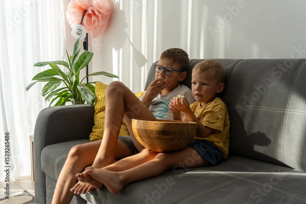 Obraz Two young brothers sitting on cozy sofa in sunny living room eating popcorn from big wooden bowl and watching movie with serious faces. Concept of childhood, bonding, home comfort and entertainment.