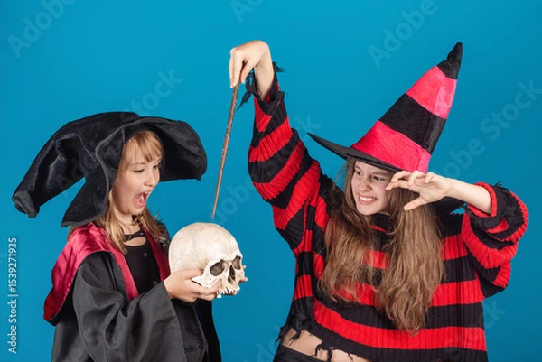 Obraz Halloween Theme. Young witch girl in striped sweater and black hat holding magic stick and making spell to the skull in another young sorceress hands against blue background