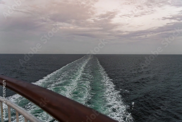 Fototapeta A tranquil view of the wide ocean as a cruise ship creates a wake, leaving behind ripples on the water. The sky carries soft hues of twilight, adding to the serene atmosphere