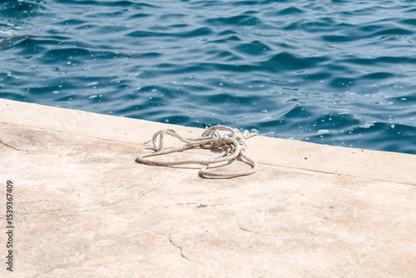 Fototapeta "a coiled rope rests on a sunlit concrete pier beside a tranquil blue ocean, symbolizing nautical themes and seaside serenity. ideal for marine and travel concepts."