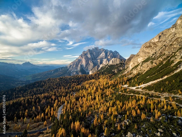 Obraz Aerial view of autumn trees and a winding road leading to a mountain range under a cloudy sky