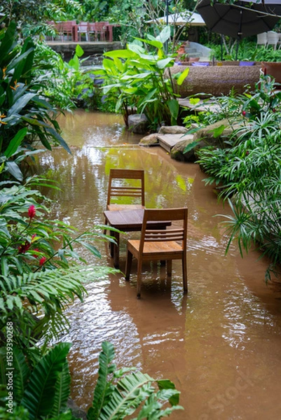 Obraz Chairs and table submerged in flooded garden, surrounded by lush greenery and tropical plants
