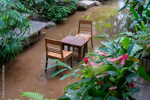 Obraz Wooden table and chairs are set in tranquil, flooded garden surrounded by lush greenery