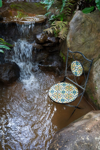 Obraz Decorative chair with colorful mosaic tiles sits in serene garden setting by small waterfall
