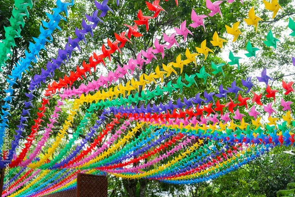 Obraz Colorful paper pinwheels hanging in rows create vibrant canopy against backdrop of green trees