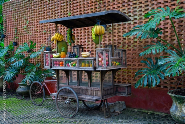 Obraz Rustic fruit cart with bananas, pineapples, and apples against brick wall, surrounded by lush green plants