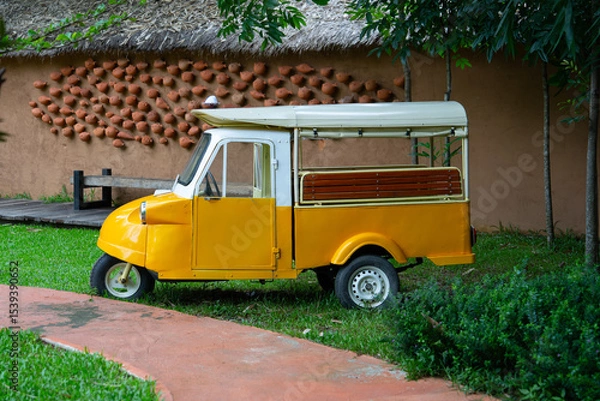 Obraz Vintage yellow tuk tuk parked on grass near rustic wall, evoking nostalgic and charming atmosphere