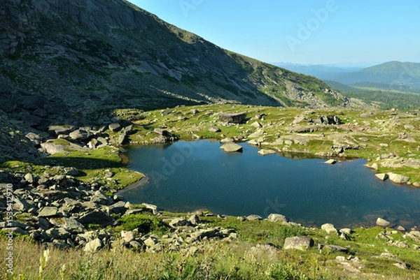 Obraz A small picturesque lake with rocks scattered along the banks at the foot of a high cliff on a sunny summer morning.
