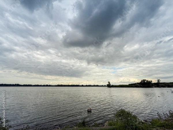 Fototapeta Wide angle view across the Danube at Grocka, a Belgrade suburb in Serbia: calm river surface under brooding summer clouds, low wooded banks and a small island form the distant horizon.