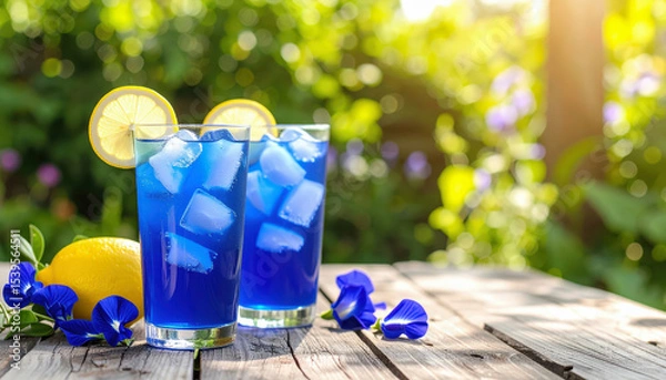 Fototapeta Blue butterfly pea flower iced tea with lemon slices on wooden table in sunny garden. Refreshing and vibrant
