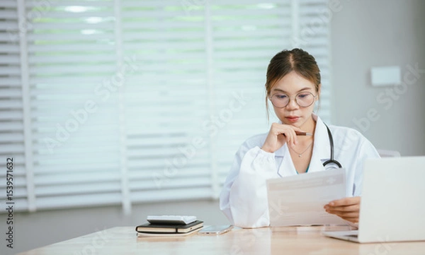 Obraz A concerned female doctor in a white lab coat and stethoscope holds her forehead, reading a document at her desk with a laptop.