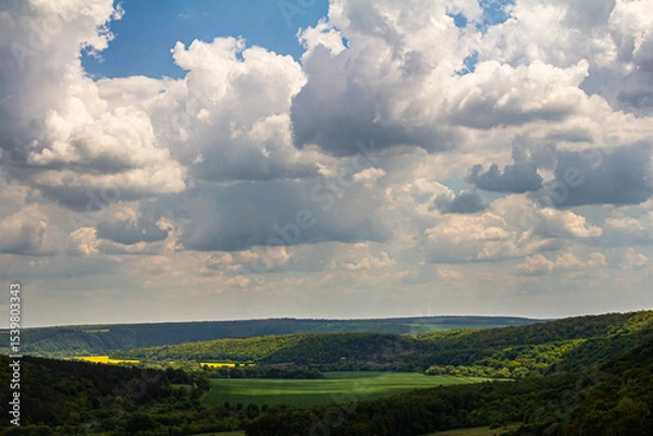 Obraz Scenic view of rolling hills and dramatic clouds over a lush green valley under a bright blue sky during midday