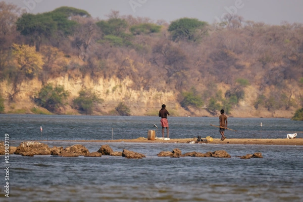 Obraz Fishermen stand on a sandbank in the Kavango River, between Namibia and Angola, in the Caprivi Strip, surrounded by serene waters and distant forested cliffs