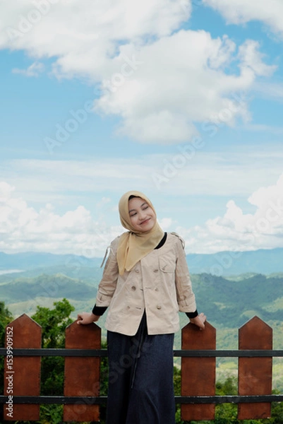 Fototapeta Smiling woman in hijab poses by a fence overlooking a scenic mountain range and cloudy sky.