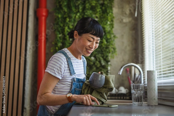 Obraz Japanese woman replies to messages while wiping dishes at home kitchen