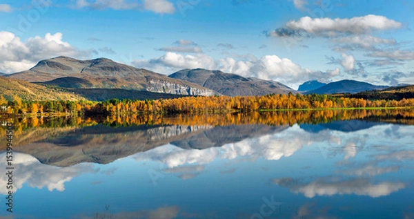 Fototapeta beautiful scenic landscape with mountain and autumnal forest under the sky reflecting in surface of water in Norway.