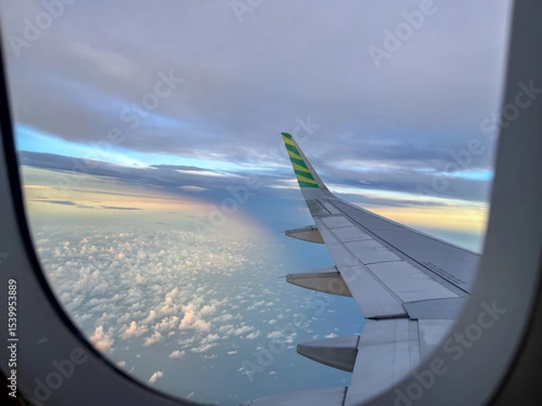 Fototapeta Wing view from an airplane window during flight, showcasing a serene sky with scattered clouds below and a soft rainbow glow on the horizon at golden hour.
