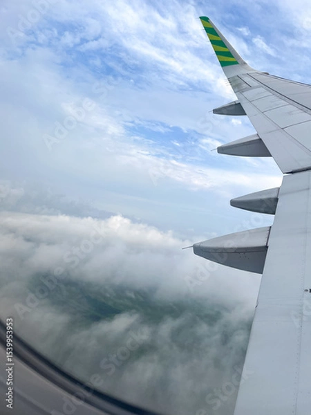 Fototapeta Airplane wing soars above fluffy clouds with patches of green landscape visible below, set against a bright blue sky in daylight, viewed from the window seat.