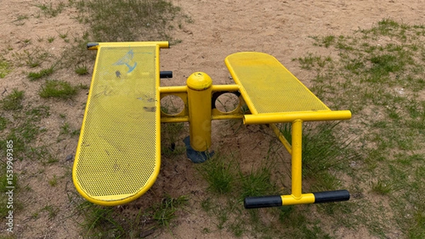 Obraz Outdoor yellow metal fitness bench for leg and abdominal workouts in a sandy playground with grass patches