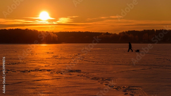 Fototapeta Panoramic Winter View of a Frozen Forest Lake with a Fisherman Walking Toward the Sunrise