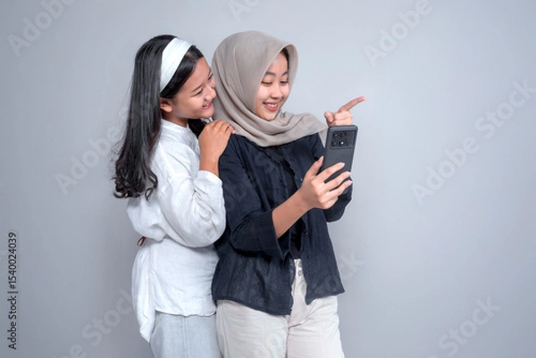 Fototapeta Two young Asian women smiling while taking a selfie. One wears a hijab and points at the phone screen. They appear happy, friendly, and close, standing in front of a plain background.