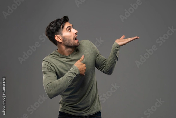Fototapeta Surprised shocked man pointing and presenting with open hand against isolated gray background studio shot