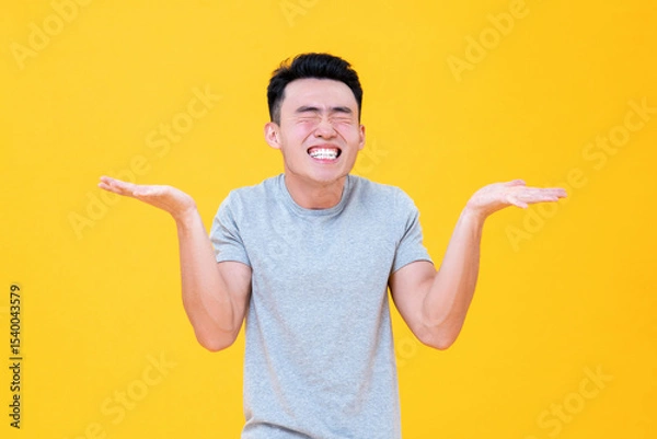Fototapeta Carefree Asian man smiling with eyes closed and open hands shrugging on yellow background
