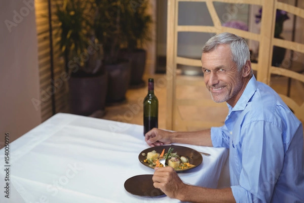 Fototapeta Senior man holding fork and knife at restaurant table with plated meal and wine, copy space