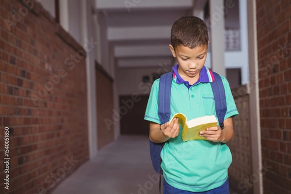 Fototapeta Boy standing in school corridor with brick walls reading yellow book and wearing blue backpack